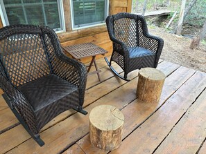 Deux chaises en osier avec un dossier et une assise tissés, une petite table carrée et deux bouts de bois sur une terrasse en bois.