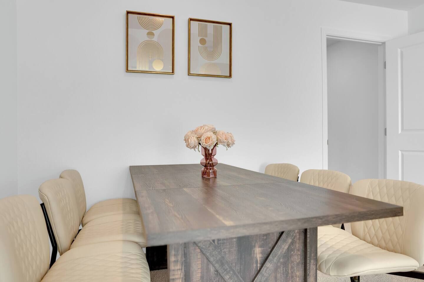 Uma sala de jantar com uma mesa de madeira, seis cadeiras estofadas e duas obras de arte emolduradas na parede.