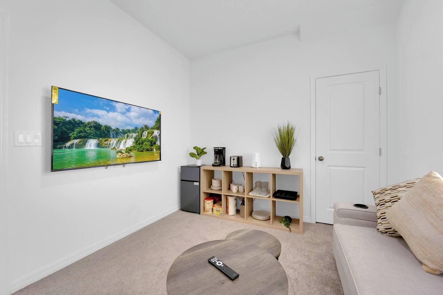 A living room with a flat-screen TV mounted on the wall, a small coffee table, a sofa, and a cabinet with various items.