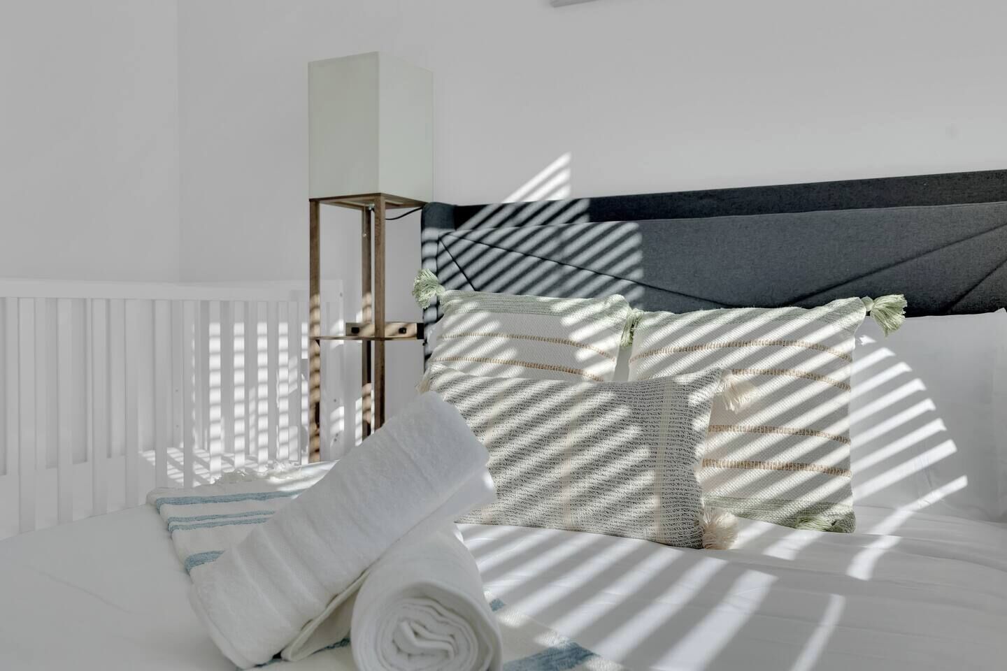 Quarto moderno com um berço branco, uma mesa de cabeceira com abajur e uma cabeceira com painel escuro.