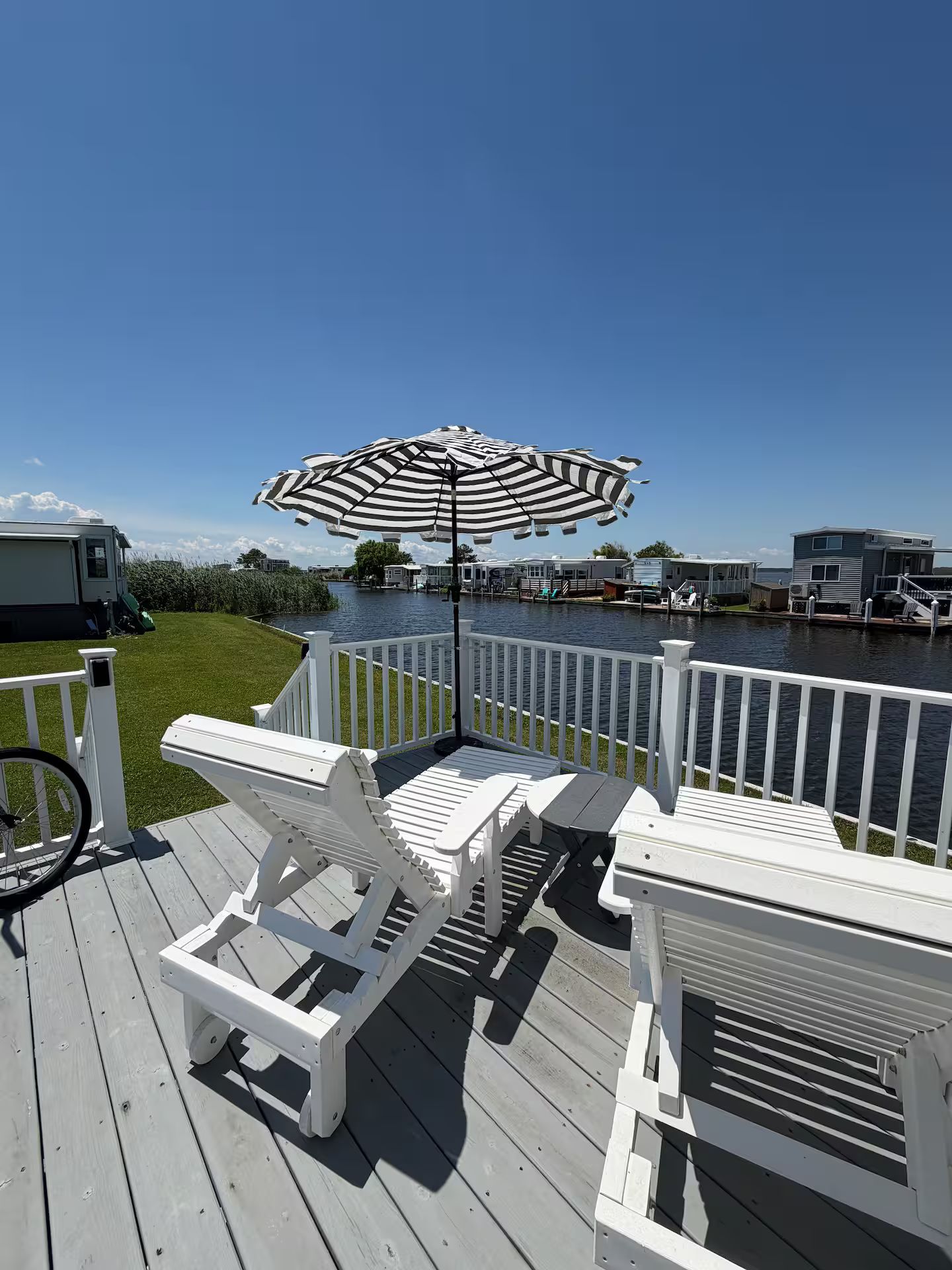 A deck with white lounge chairs and a striped umbrella, overlooking a body of water with mobile homes in the background.