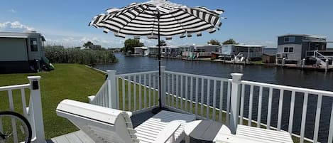 A deck with white lounge chairs and a striped umbrella, overlooking a body of water with mobile homes in the background.