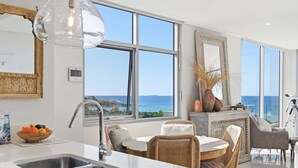 A modern kitchen with a large window, a dining area with wicker furniture, and a view of the sea. - Kai - Penthouse Luxury with breathtaking Ocean views (Coffs Harbour)