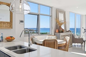 Eine moderne Küche mit einem großen Fenster, ein Essbereich mit Korbmöbeln und Blick aufs Meer.