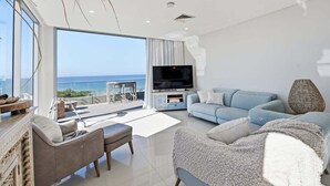 A modern living room with a large TV, a sofa, and a balcony with a view of the sea. - Kai - Penthouse Luxury with breathtaking Ocean views (Coffs Harbour)