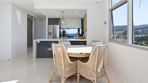 A modern kitchen with a view, a dining area with wicker chairs, and a large window. - Kai - Penthouse Luxury with breathtaking Ocean views (Coffs Harbour)