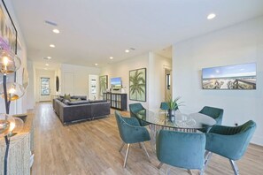 A modern living room with a glass dining table, teal chairs, a gray sofa, and a large framed picture on the wall. - Salt Life: Private Beach, Gulf Views, Heated Pool & Spa, Golf Cart, 7,700+ SqFt (Miramar Beach)