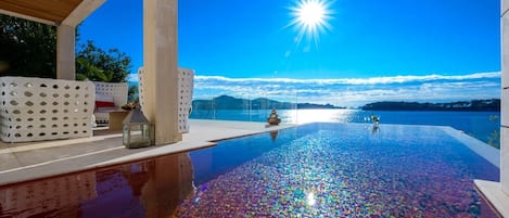 An infinity pool with a clear view of the horizon and mountains, a wooden ceiling, and a white outdoor seating area.