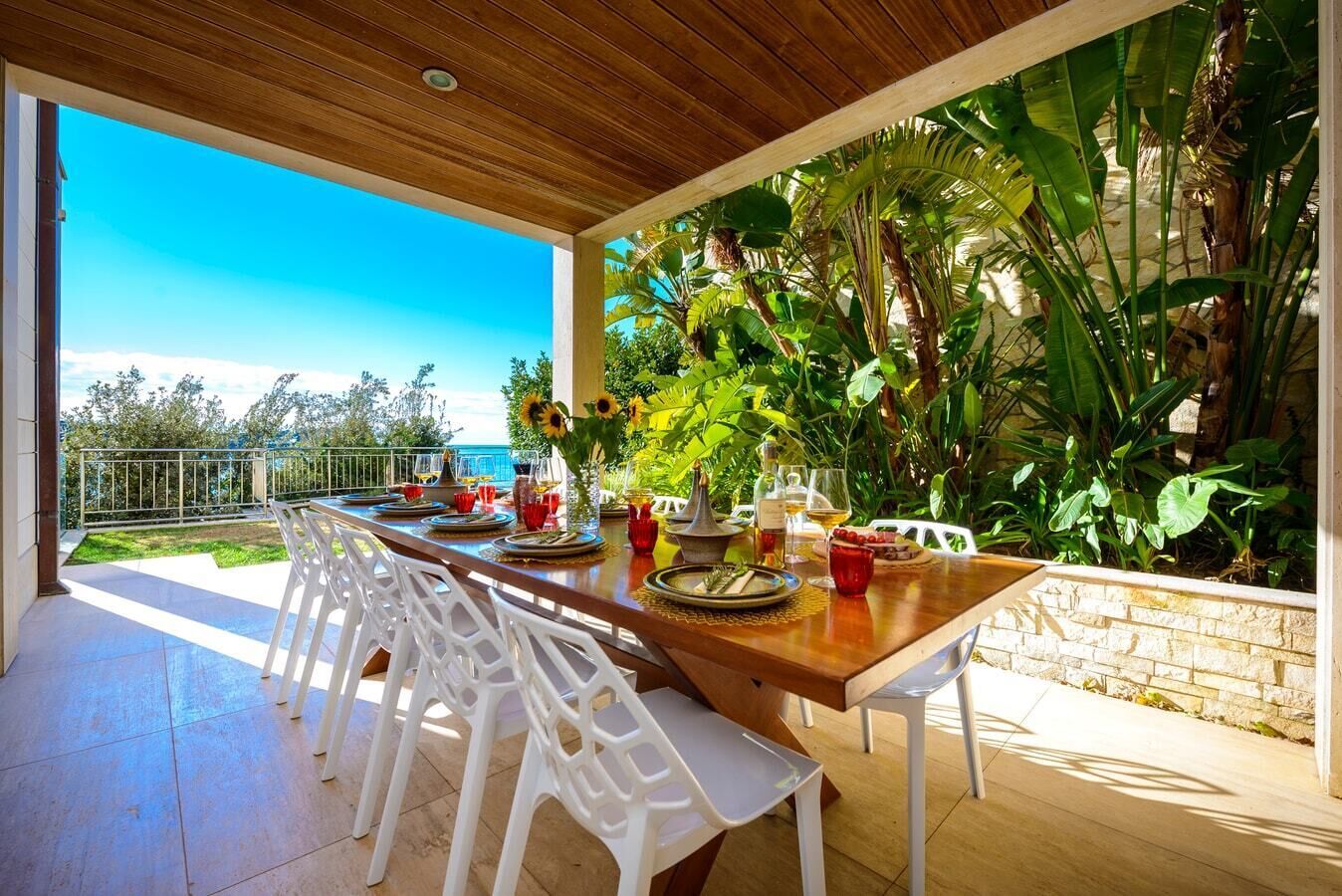 An outdoor dining area with a wooden table, white chairs, and a view of the sea.