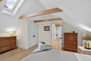Ein Schlafzimmer mit schrägem Dach, Holzbalken, einem Bett, einem Schminktisch und einer Nachttischlampe.