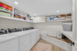 A laundry room with a row of washing machines, shelves with folded clothes, and a small heater. - Family Friendly Home Just Minutes from Boothbay Harbor! (Boothbay)