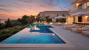 Piscina com espreguiçadeiras e guarda-sóis, rodeada por um jardim ajardinado e um edifício com varanda.
