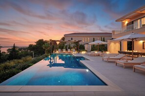Ein Swimmingpool mit Liegestühlen und Sonnenschirmen, umgeben von einem angelegten Garten und einem Gebäude mit Balkon.