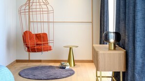Um quarto com uma cadeira suspensa, uma mesinha redonda, uma mesinha de apoio com abajur e uma cortina azul.