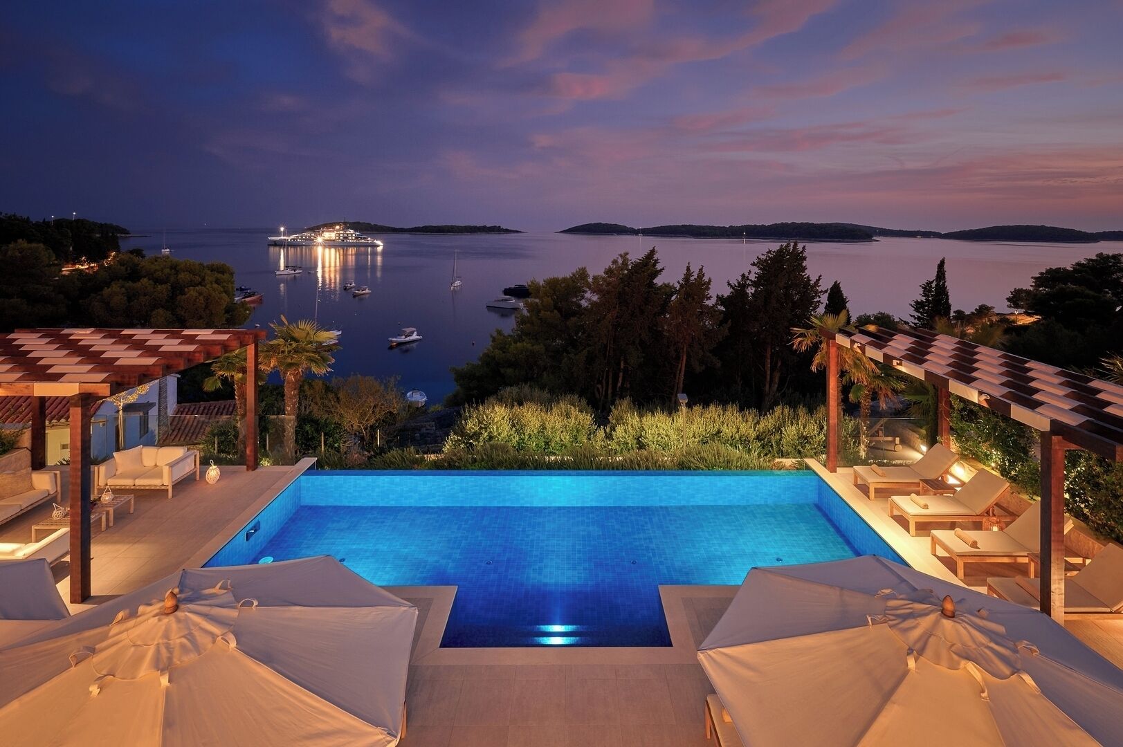 A swimming pool with lounge chairs and umbrellas, surrounded by greenery and overlooking a body of water with boats.
