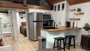 A modern kitchen with a stainless steel refrigerator, a microwave, and wooden countertops. There are two black bar stools and a round table with a basket containing a plant and a remote control.