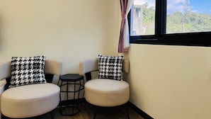 Une pièce d’angle avec deux chaises et une petite table, une fenêtre avec des rideaux et une vue sur un immeuble et des arbres.