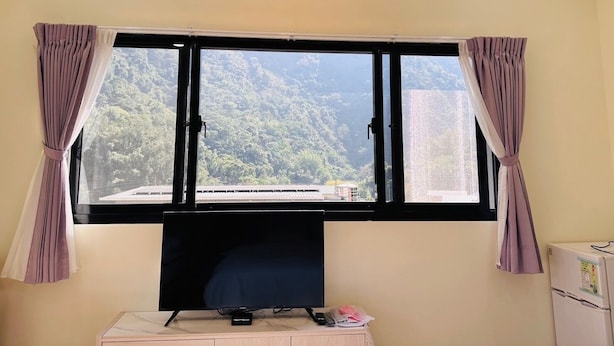 Une chambre dotée d’une grande fenêtre, d’un téléviseur à écran plat, d’une petite table avec une télécommande et d’un réfrigérateur.