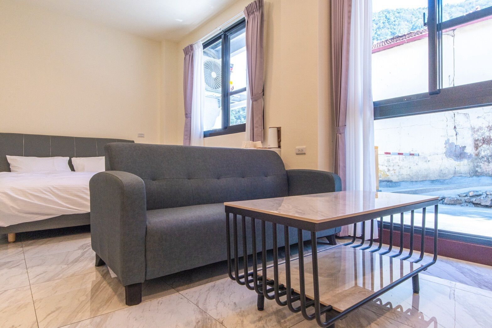 A room with a bed, a grey sofa, a small table, and a window with curtains.