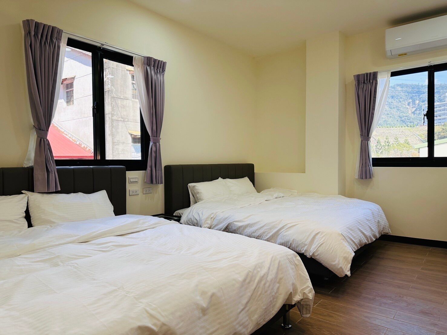 A hotel room with two beds, a window with curtains, an air conditioning unit, and a view of buildings outside.