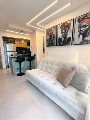 A modern living room with a white sofa, a kitchen in the background, and three framed artworks on the wall. - Apartamento 3 hab. con piscina Cerca a la playa. (Cartagena de Indias)