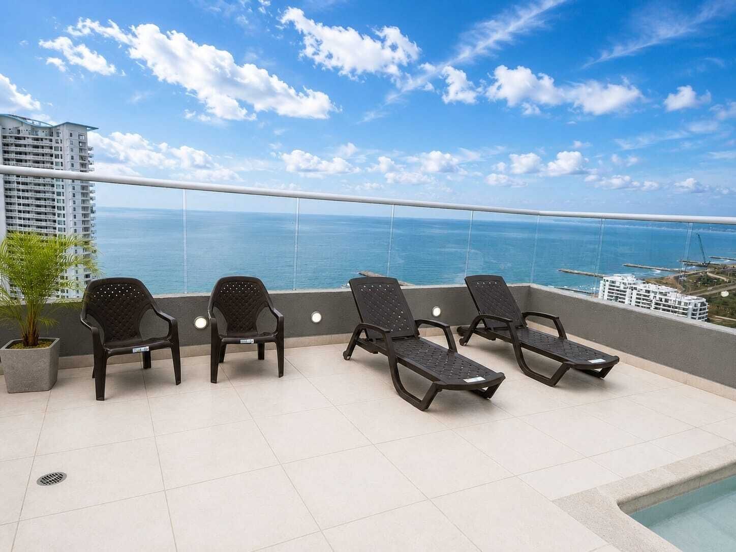 Ein Balkon mit vier Liegestühlen, einer Topfpflanze und Blick auf das Meer und Gebäude.