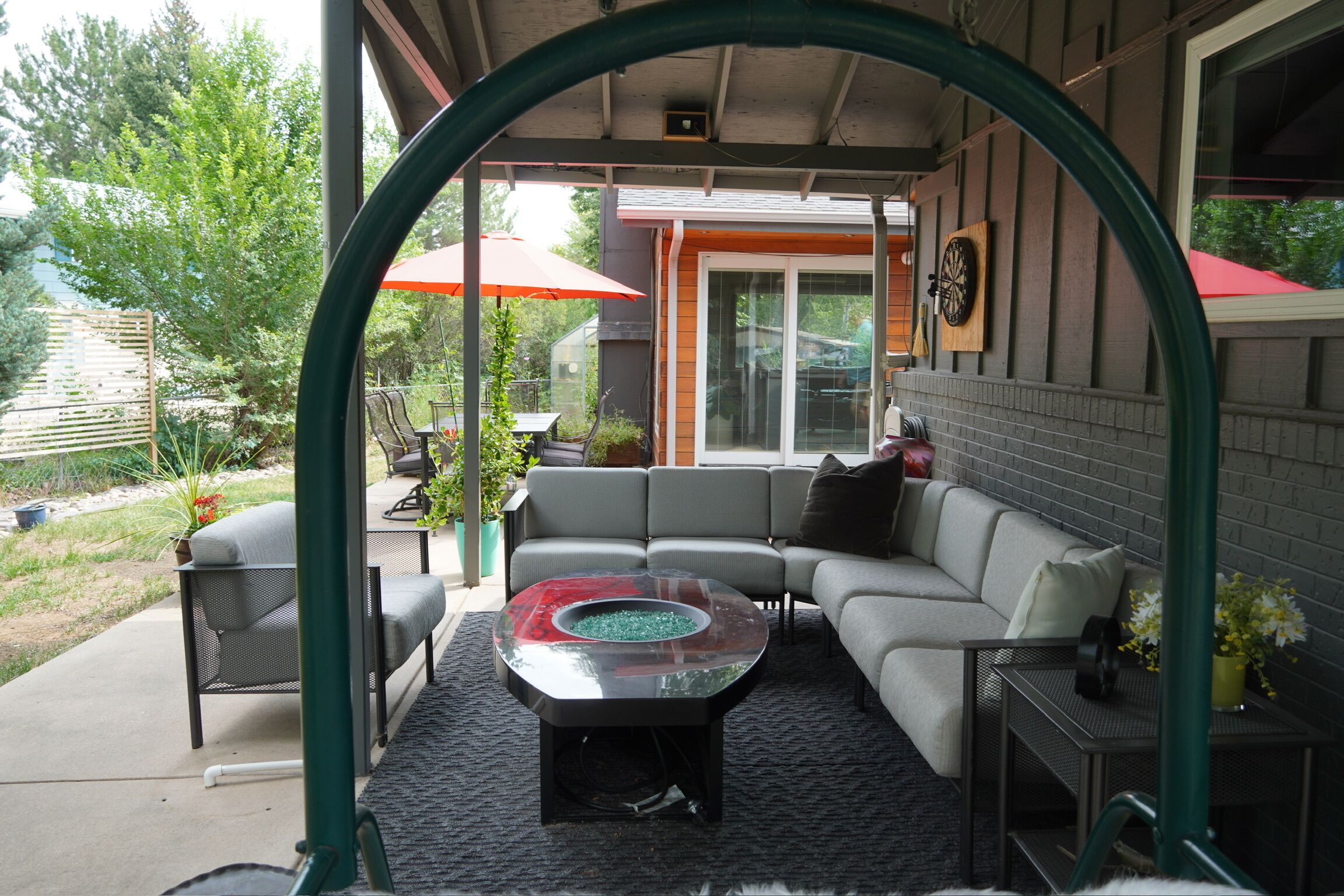 Een terras buiten met een gebogen dak, een hoekbank, een salontafel met glazen blad en een rode parasol.