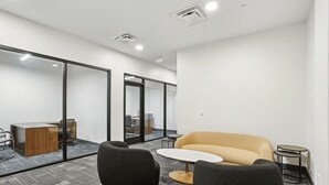 Een moderne kantoorruimte met een glazen wand, een bank, twee stoelen, een klein rond tafeltje en een bureau met een computer.
