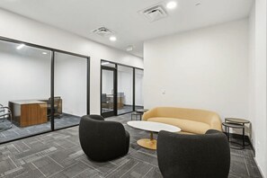 A modern office space with a glass partition, a sofa, two chairs, a small round table, and a desk with a computer. - Upscale Downtown 2BR | King + Secured Parking (Birmingham)