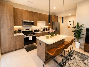 A modern kitchen with wooden cabinetry, stainless steel appliances, a central island with bar stools, and a potted plant. - Upscale Downtown 2BR | King + Secured Parking (Birmingham)