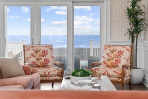 Habitación con dos sillones tapizados de estampado floral, un sofá, una mesa de centro y vistas al océano a través de amplias ventanas.