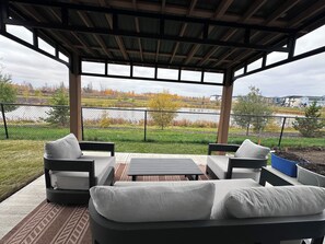 Une terrasse extérieure dotée d’un espace salon couvert, offrant une vue sur un lac et entourée d’une clôture.