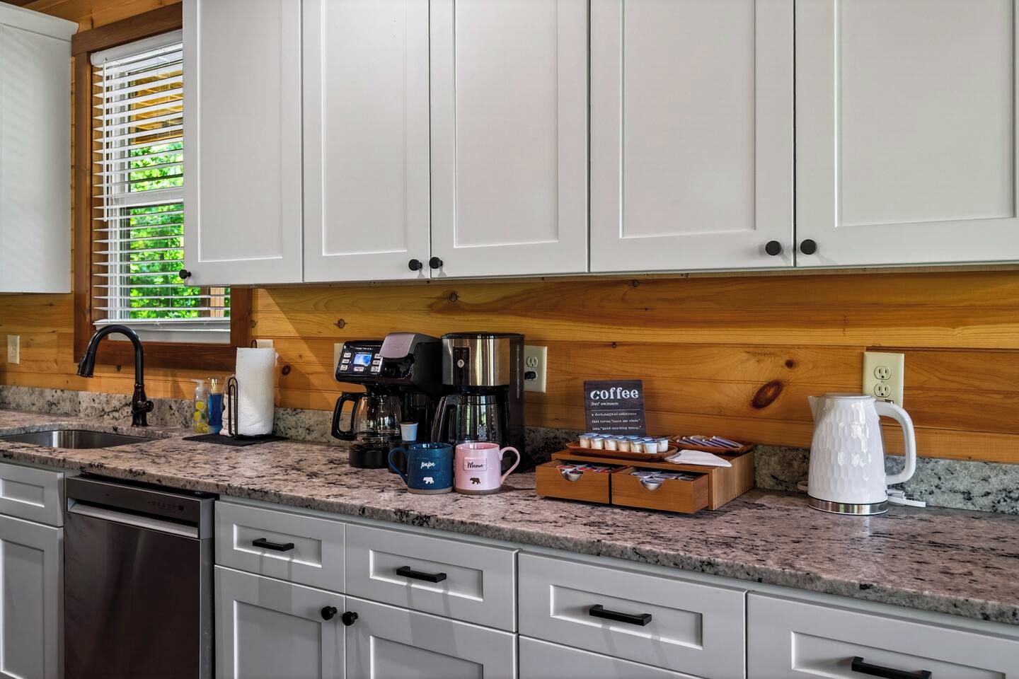 A kitchen with white cabinets, a granite countertop, a coffee maker, mugs, and a kettle.