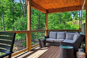 Ein überdachter Außenbereich mit Korbmöbel-Sofa und Bank, umgeben von einer Holzterrasse und einem Drahtzaun.