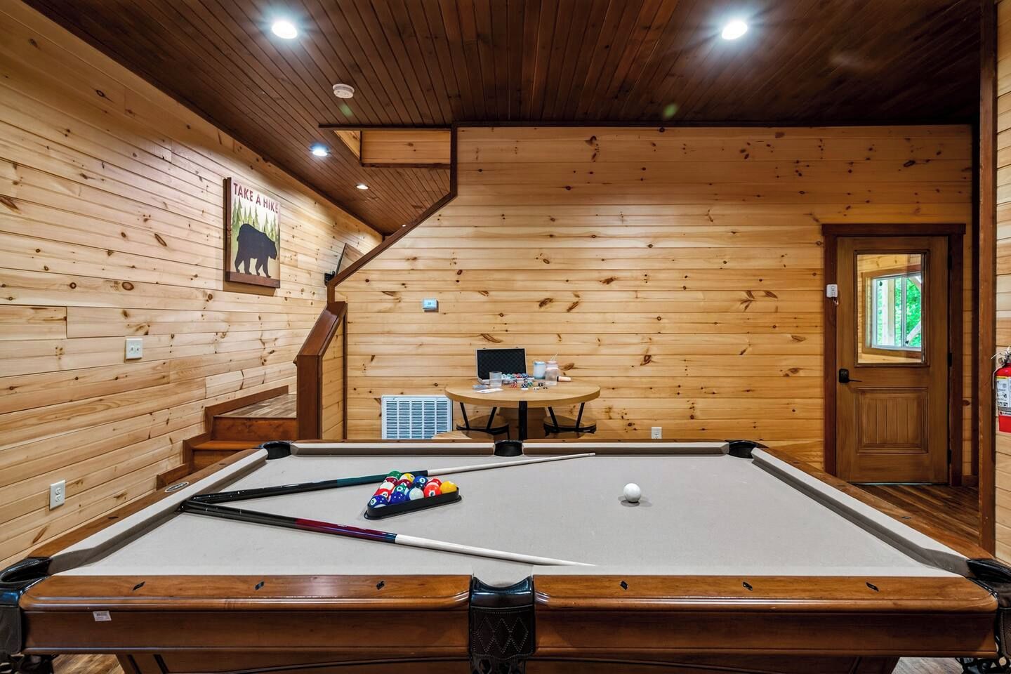 Una mesa de billar con bolas, una computadora y una pequeña mesa con objetos encima, todo en una habitación con paredes de madera y una puerta.