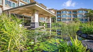 Un hotel con piscina rodeada de nenúfares y plantas tropicales, una entrada cubierta y varios balcones.