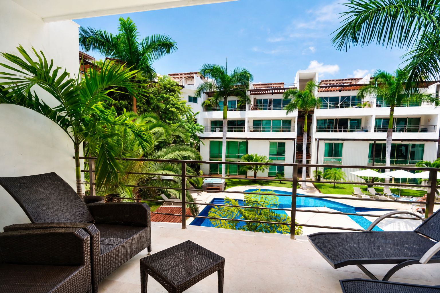 A balcony with wicker furniture overlooking a swimming pool and a building with multiple windows.
