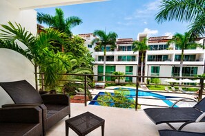 A balcony with wicker furniture overlooking a swimming pool and a building with multiple windows. - Playa Paradise 4BR & 4BA Penthouse + Private Rooftop + 5 Min Walk to Beach (Playa del Carmen)