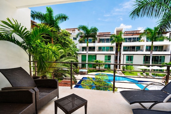 A balcony with wicker furniture overlooking a swimming pool and a building with multiple windows.