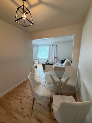 Ein modernes Wohnzimmer mit Esstisch aus Glas, weißen Stühlen, einer Couch und einer Deckenleuchte.