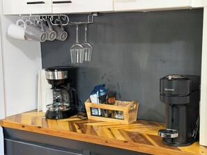 Eine Küchenarbeitsplatte mit Kaffeemaschine, einer Glaskaraffe, einer Holzkiste mit Kaffeekapseln und einem Hängegestell für Tassen und Gläser.