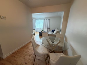 Ein Essbereich mit einem Glastisch und weißen Stühlen, ein Sofa im Hintergrund und eine weiße Decke.