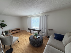 Ein modernes Wohnzimmer mit Holzboden, einem grauen Sofa, einem gemusterten Hocker, einer Bank, einem kleinen Tisch mit Kerze und einem Blumentopf.