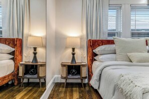 A hotel room with a bed, two bedside tables with lamps, a window with blinds, and a wooden floor. - Luxury Designer Suite 2BR• Boston • Sunroom (Boston)