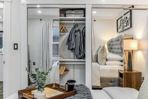 A modern bedroom with a bed, bedside table, lamp, and a view of a closet with hanging clothes. - Luxury Designer Suite 2BR• Boston • Sunroom (Boston)