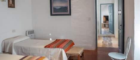 A room with two beds, a bench, a chair, a wooden ceiling, and a door leading to another room.