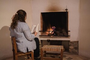 Una persona sentada frente a una chimenea, con un libro, una copa de vino y una mesita de madera.