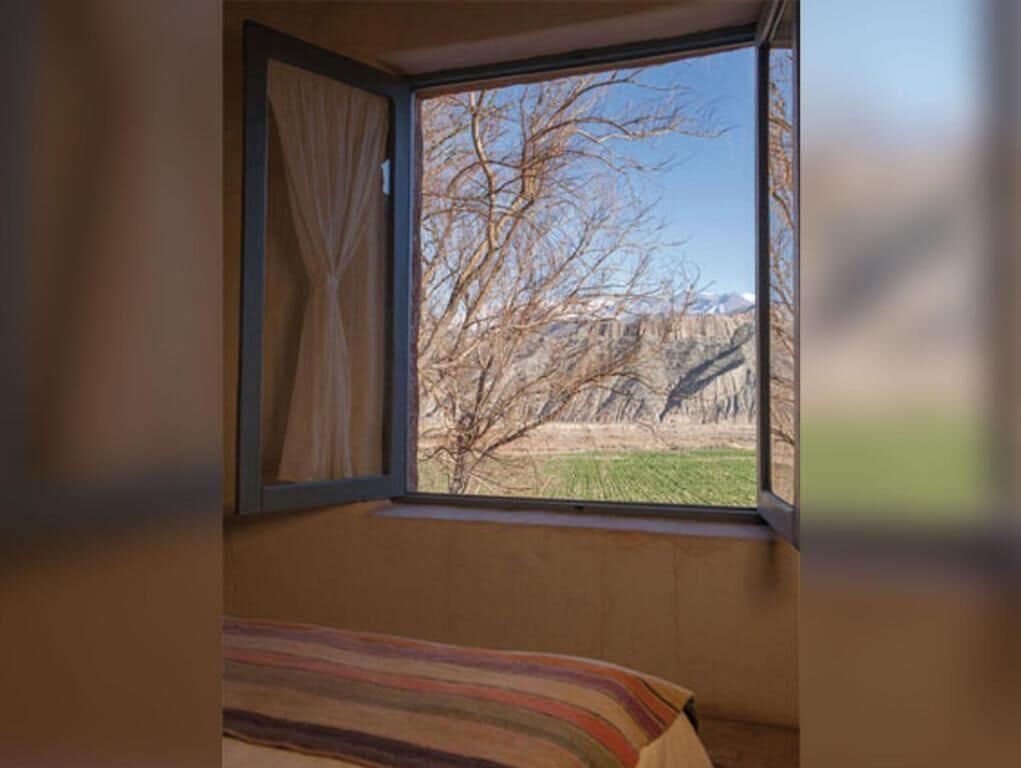 Um quarto com uma cama, uma janela com cortinas e vista para uma paisagem com árvores e montanhas.