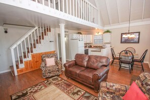 A spacious living area with a brown leather sofa, patterned armchairs, a dining table with chairs, a kitchen in the background, and a white staircase. - Viewcatcher: Gatlinburg 2BR A-Frame Cabin w/ Mountain Views, Hot Tub & Resort Pools (Gatlinburg)