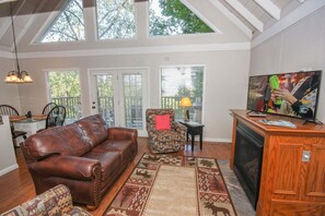 A living room with a leather sofa, patterned armchair, and a television on a wooden stand. - Viewcatcher: Gatlinburg 2BR A-Frame Cabin w/ Mountain Views, Hot Tub & Resort Pools (Gatlinburg)
